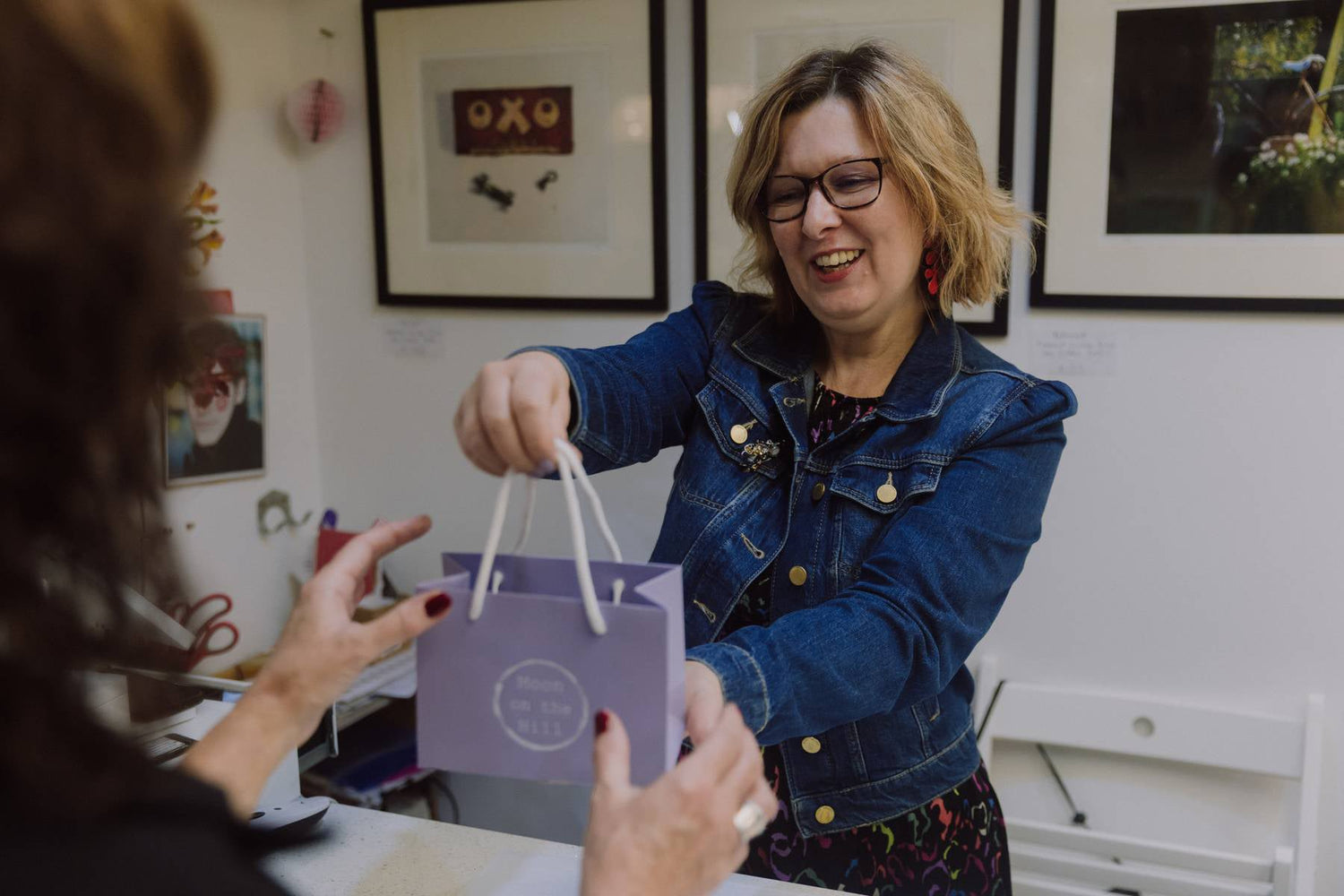Moon on the Hill business owner wearing a denim jacket and black dress, handing a customer a branded bag.